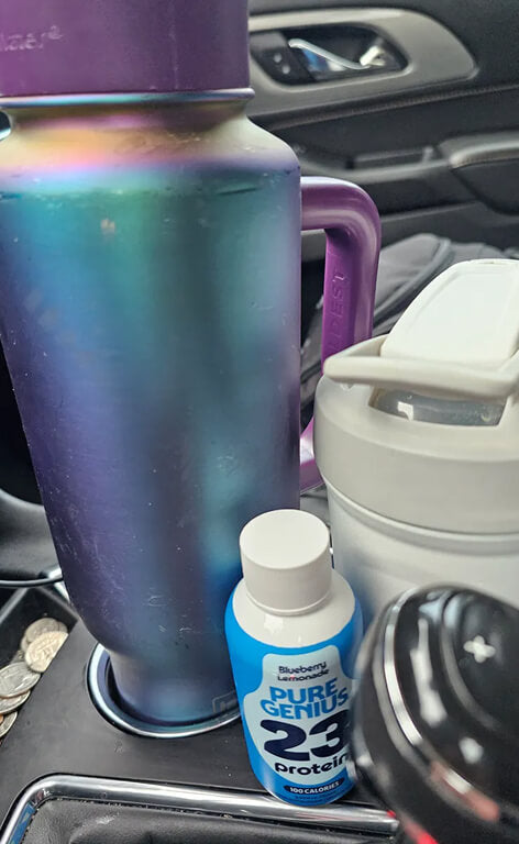 Pure Genius Blueberry Lemonade protein shot in a car cupholder beside a travel tumbler